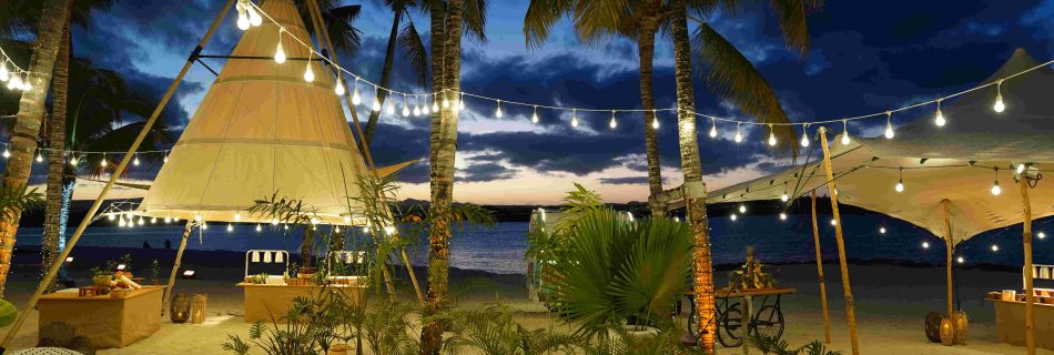Tropical beach setting at dusk with string lights hanging between palm trees, cozy tents, and lanterns, creating a warm, inviting atmosphere by the sea.