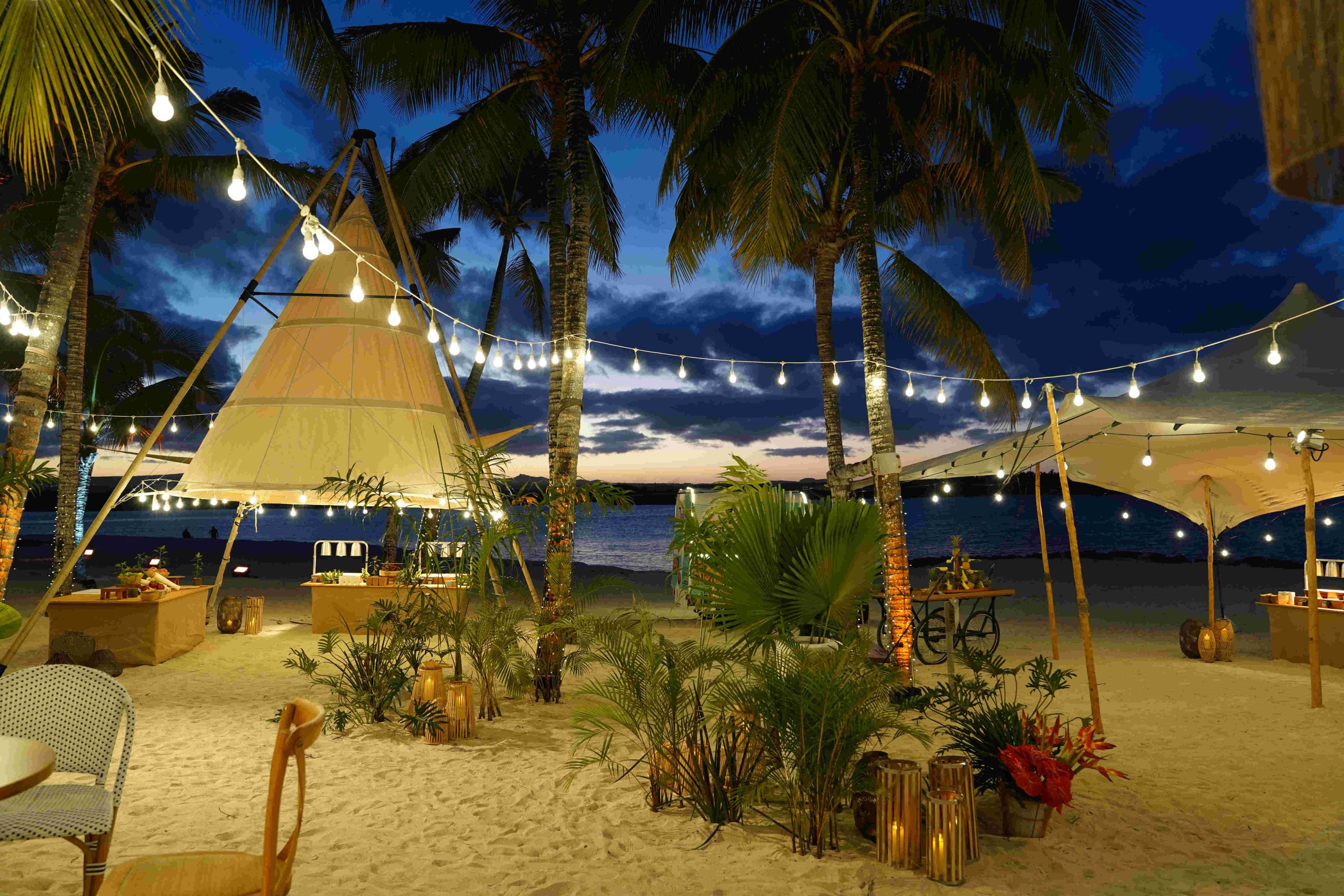 Tropical beach setting at dusk with string lights hanging between palm trees, cozy tents, and lanterns, creating a warm, inviting atmosphere by the sea.