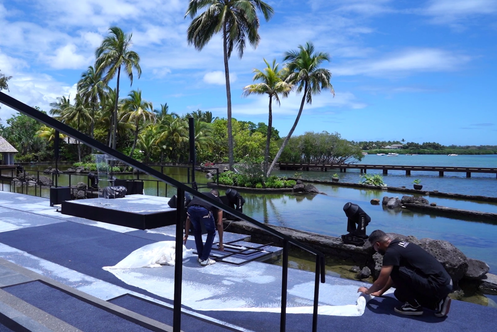 Event setup in a tropical setting with workers laying white carpets near a serene waterway. Palm trees, clear blue sky, and a tranquil ambiance.