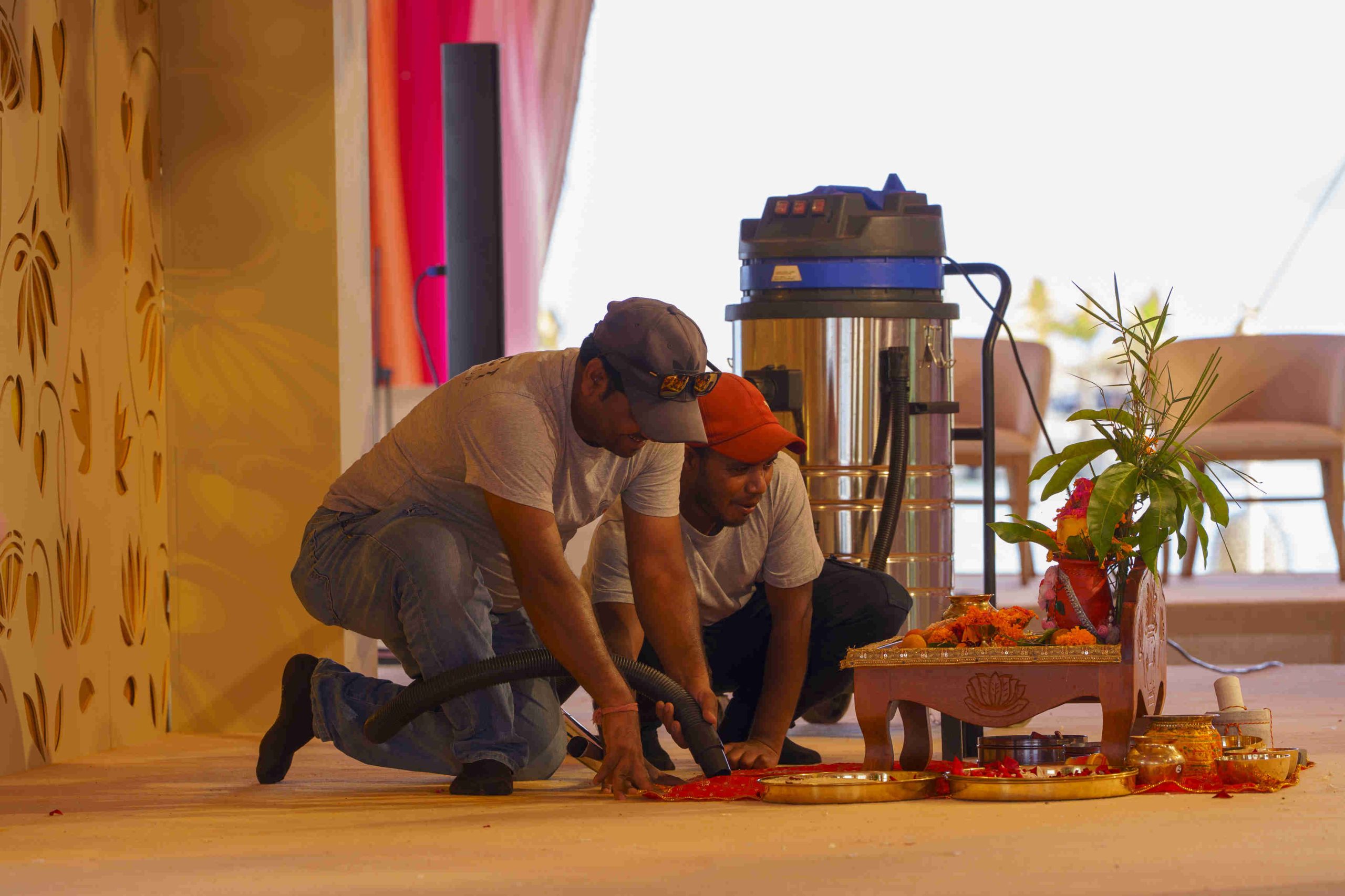 Two men kneel on a stage, using tools near a small ornate table with religious items, a plant, and vibrant decorations. A vacuum cleaner is in the background.