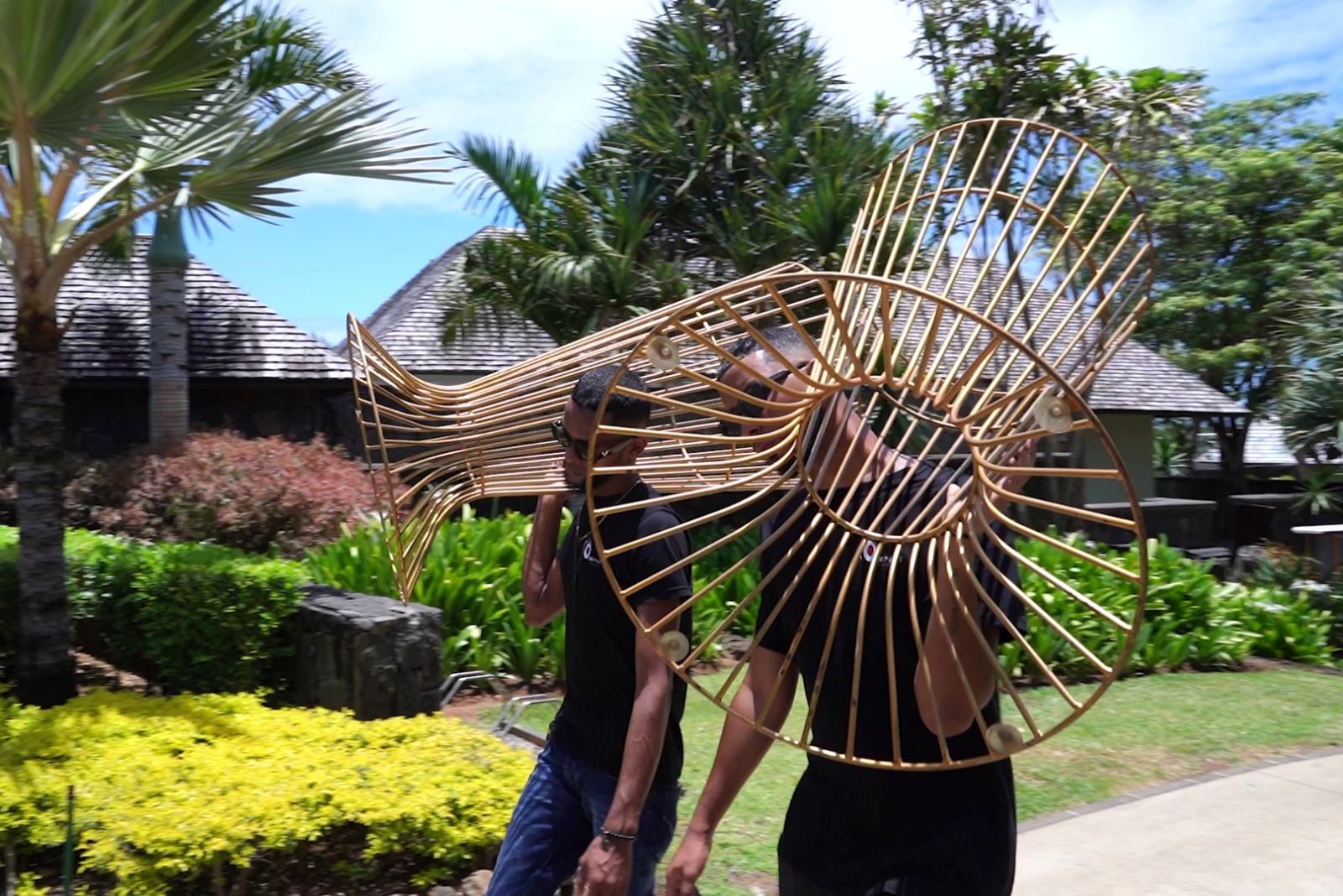 Two people carry a large wicker sculpture through a tropical garden, with lush greenery and thatched-roof buildings in the background on a sunny day.