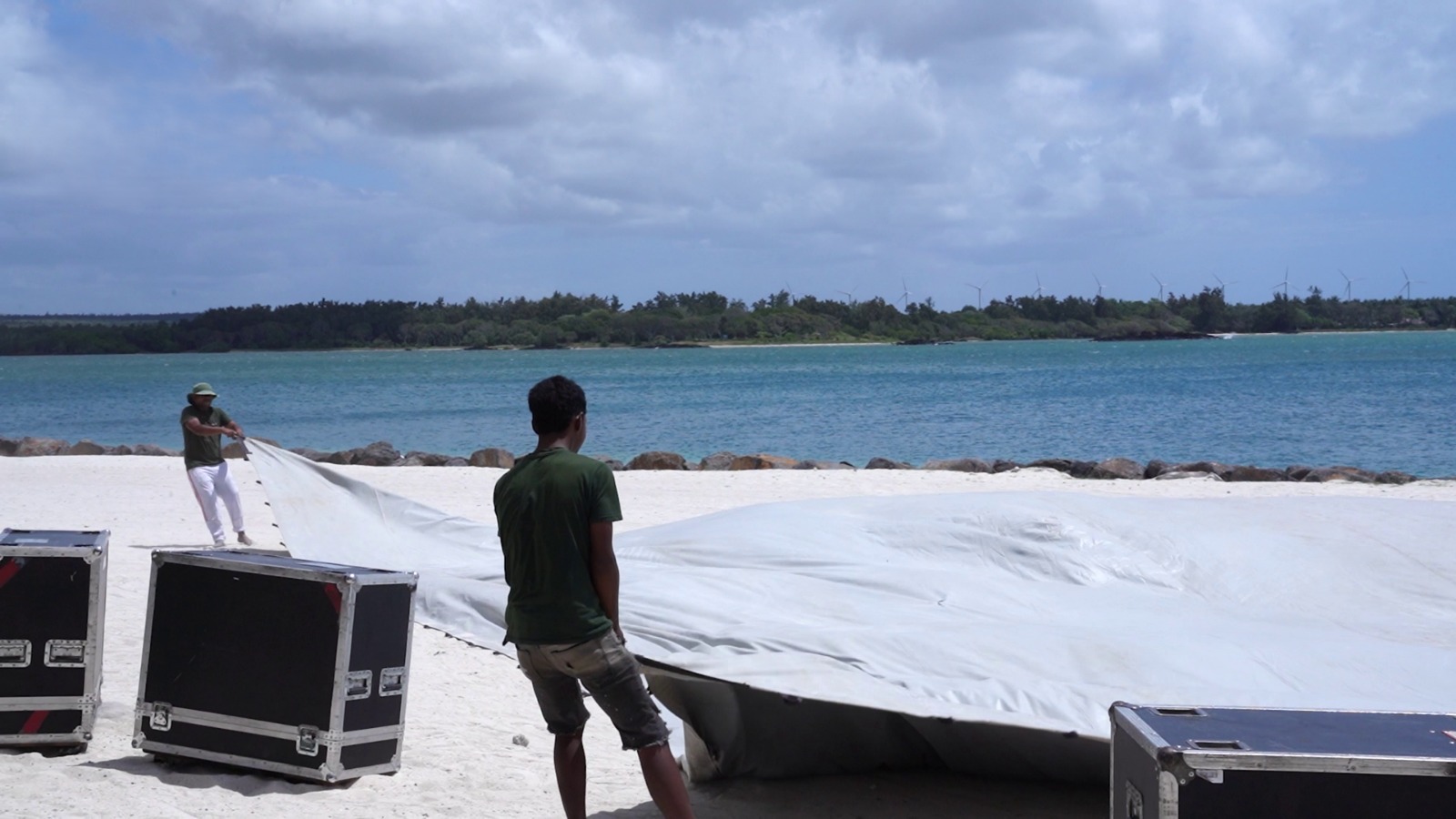Two people on a sandy beach pull a large tarp near the ocean. Equipment cases are in the foreground. The scene is calm with a cloudy sky.