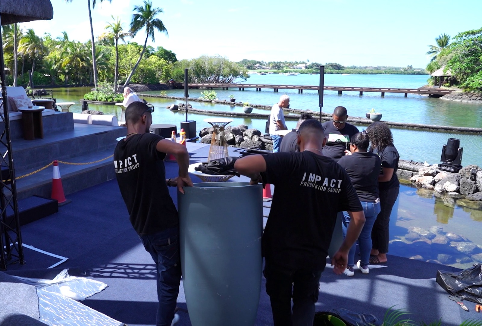 A team of people sets up by a scenic waterfront with palm trees and a clear blue sky. They're arranging decor near a modern stage.