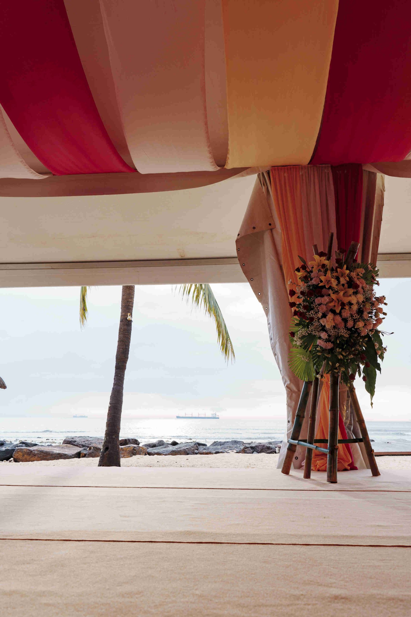 Wedding canopy with red and white drapes on a beach. A floral arrangement stands on the right, with palm trees and a ship visible in the distance. Tranquil setting.