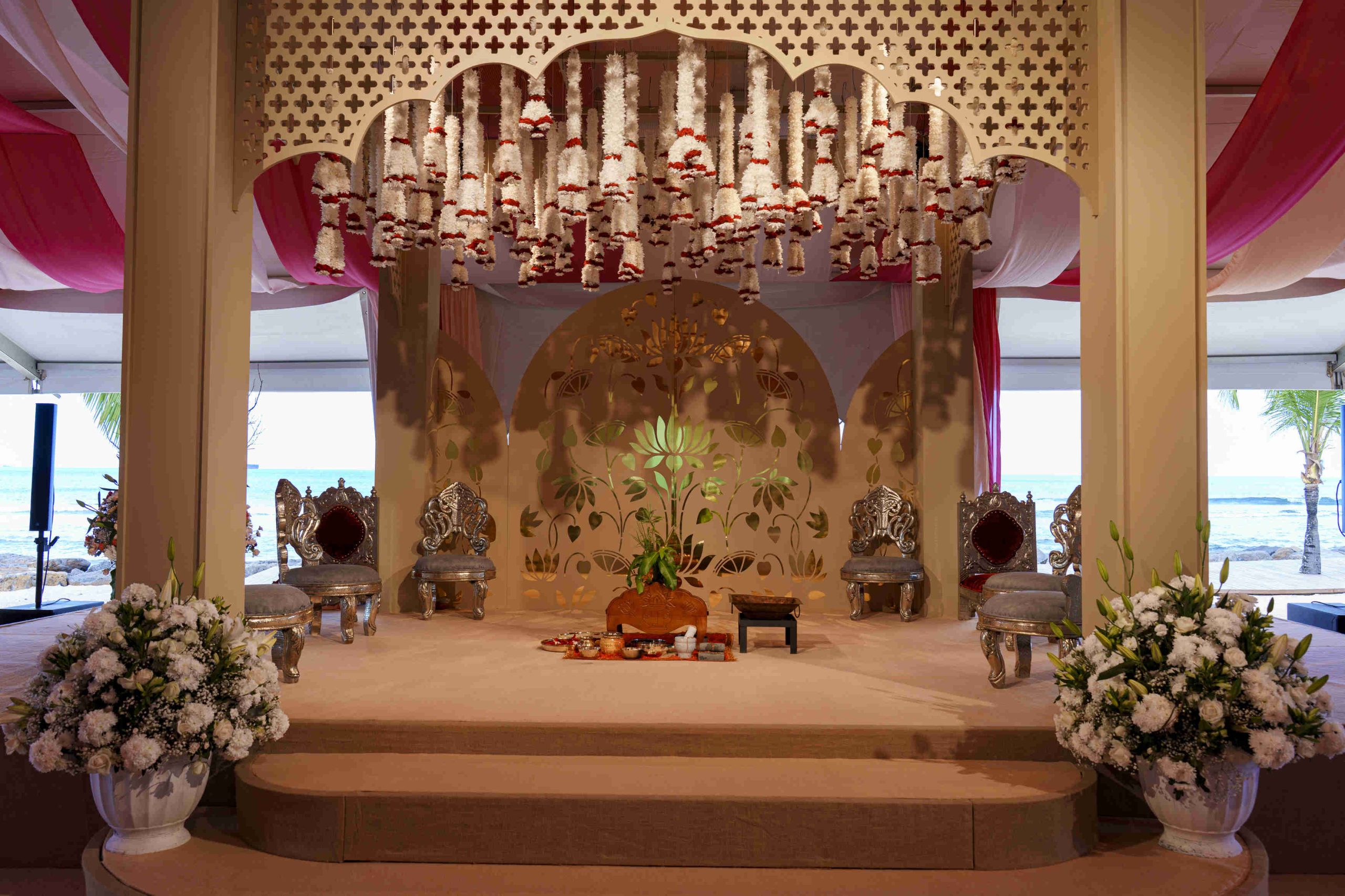 Elegant wedding mandap with ornate chairs, floral garlands, and decorative backdrop. White flowers adorn the setting, overlooking a serene beach view.