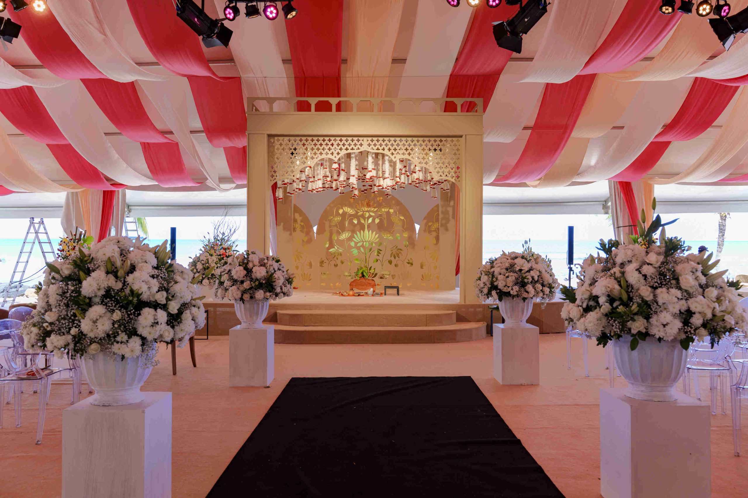 Elegant wedding stage with a gold centerpiece, surrounded by large white floral arrangements. Red and white drapes hang from the ceiling, creating a festive atmosphere.