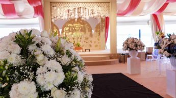 Elegant wedding venue with pink and white drapes, white floral arrangements, and a gold backdrop. The atmosphere is celebratory and stylish.