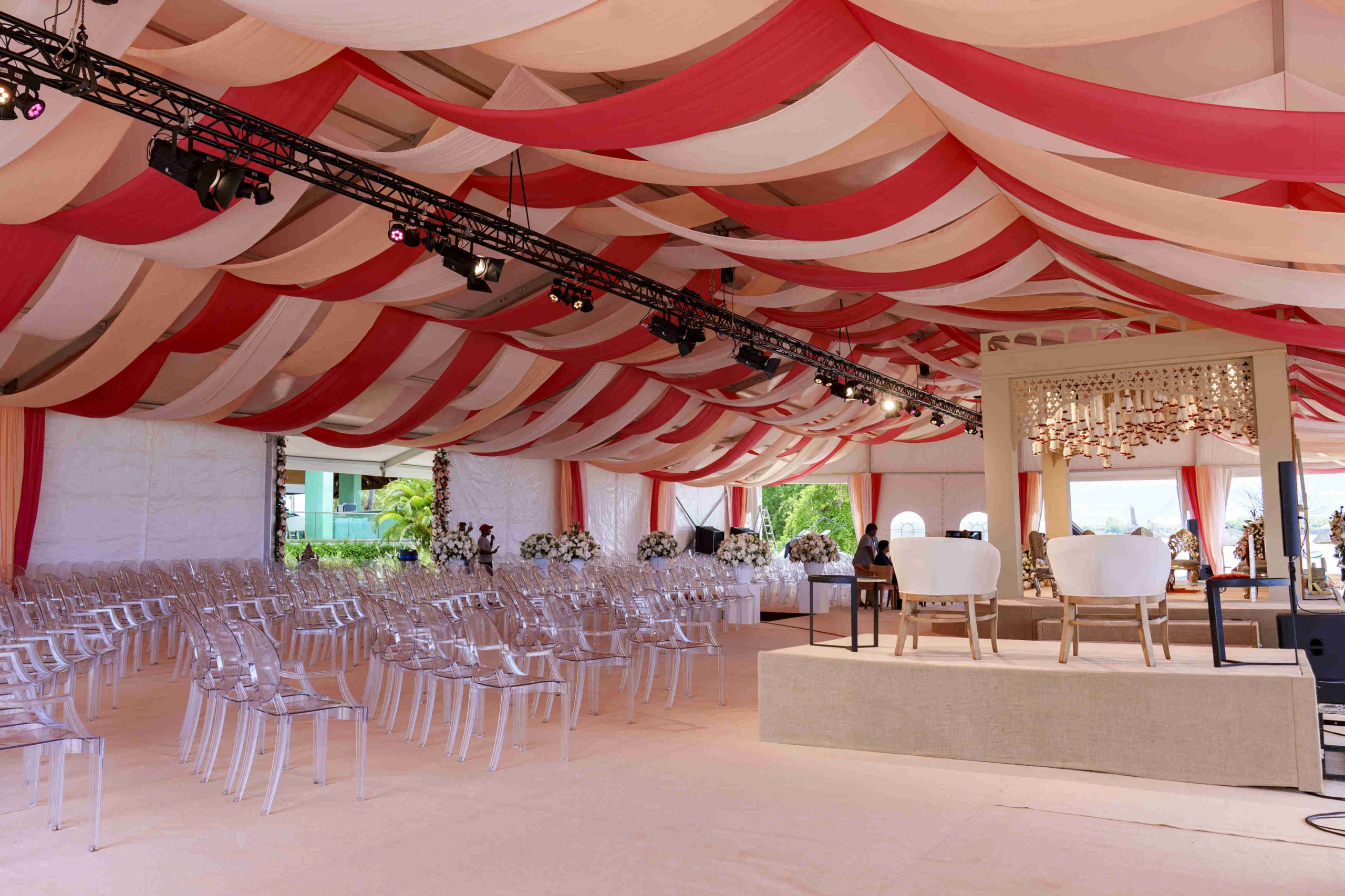 Elegant wedding venue with red and white draped ceiling, clear chairs arranged in rows, a floral-decorated stage, and a sophisticated atmosphere.