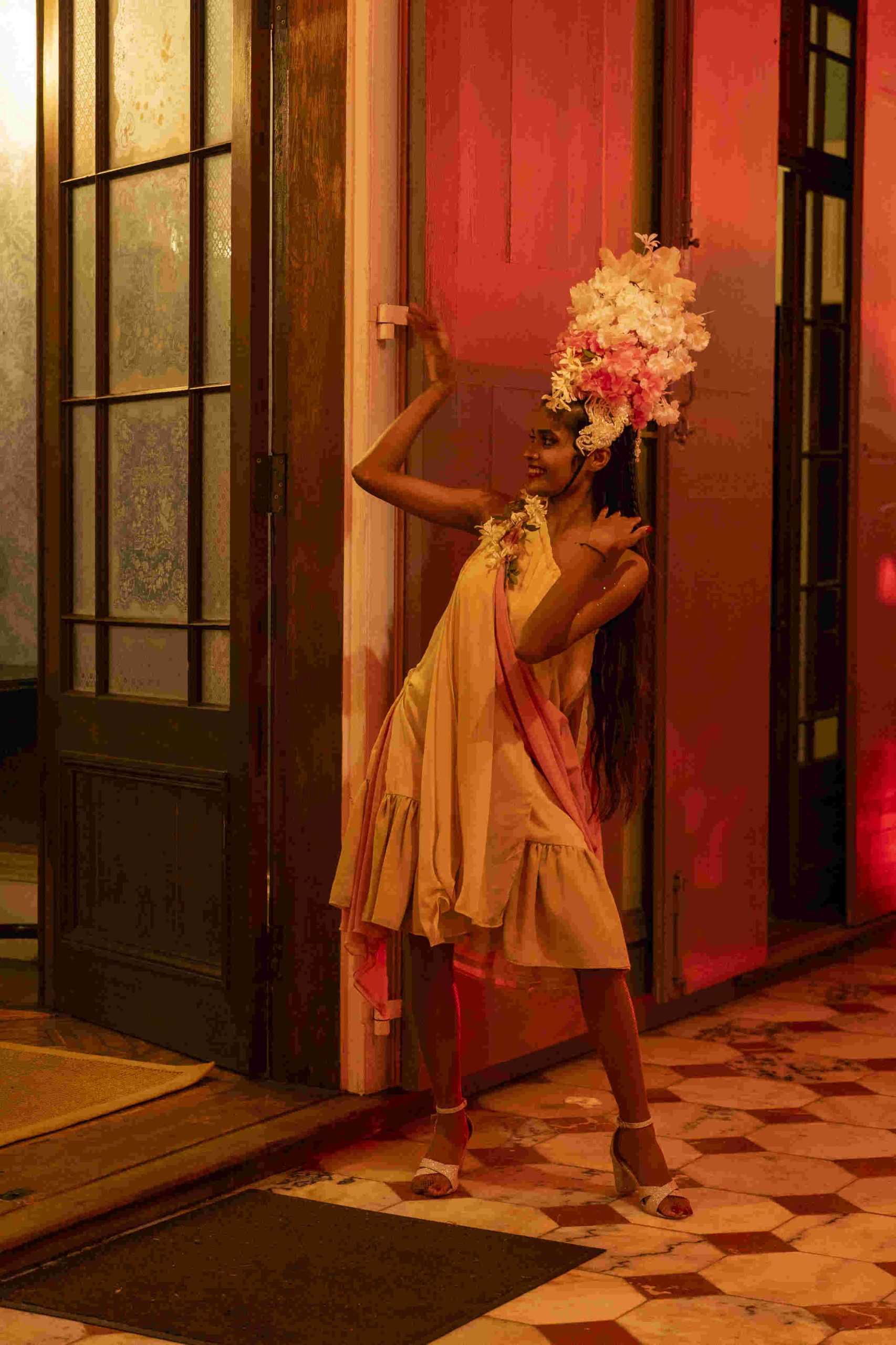 Woman in a beige dress and elaborate floral headpiece poses confidently in a warmly lit hallway. The atmosphere is artistic and lively.
