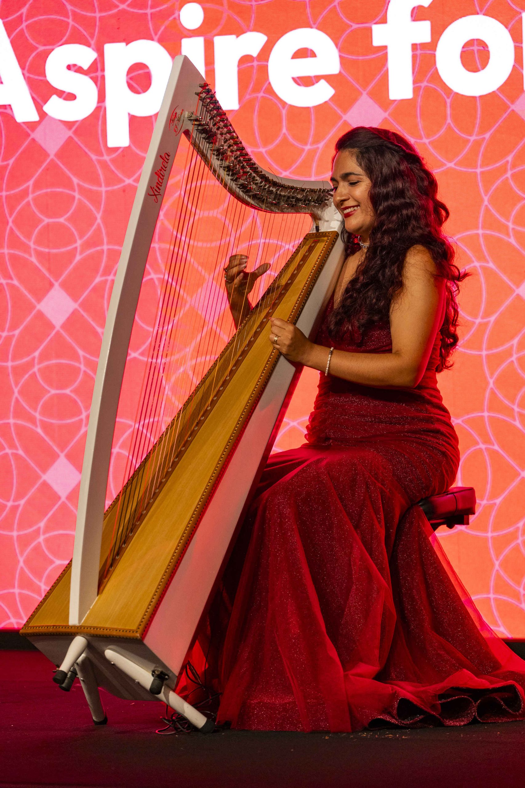 A woman in a red gown plays a large harp on stage, smiling against a red backdrop with the words "Aspire fo." The scene conveys elegance and joy.