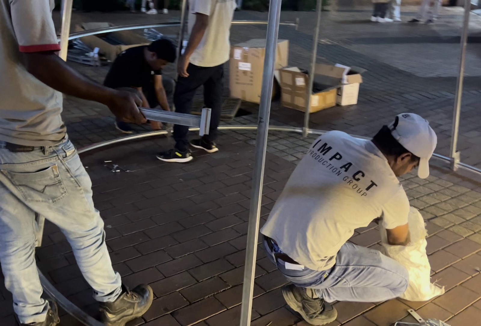 Workers in casual attire construct a framework on a brick surface, surrounded by boxes. The scene conveys teamwork and focus on task.