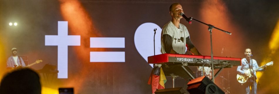 Musician playing keyboard and singing on stage with a band, backed by a large 'Cross = Heart' light display.