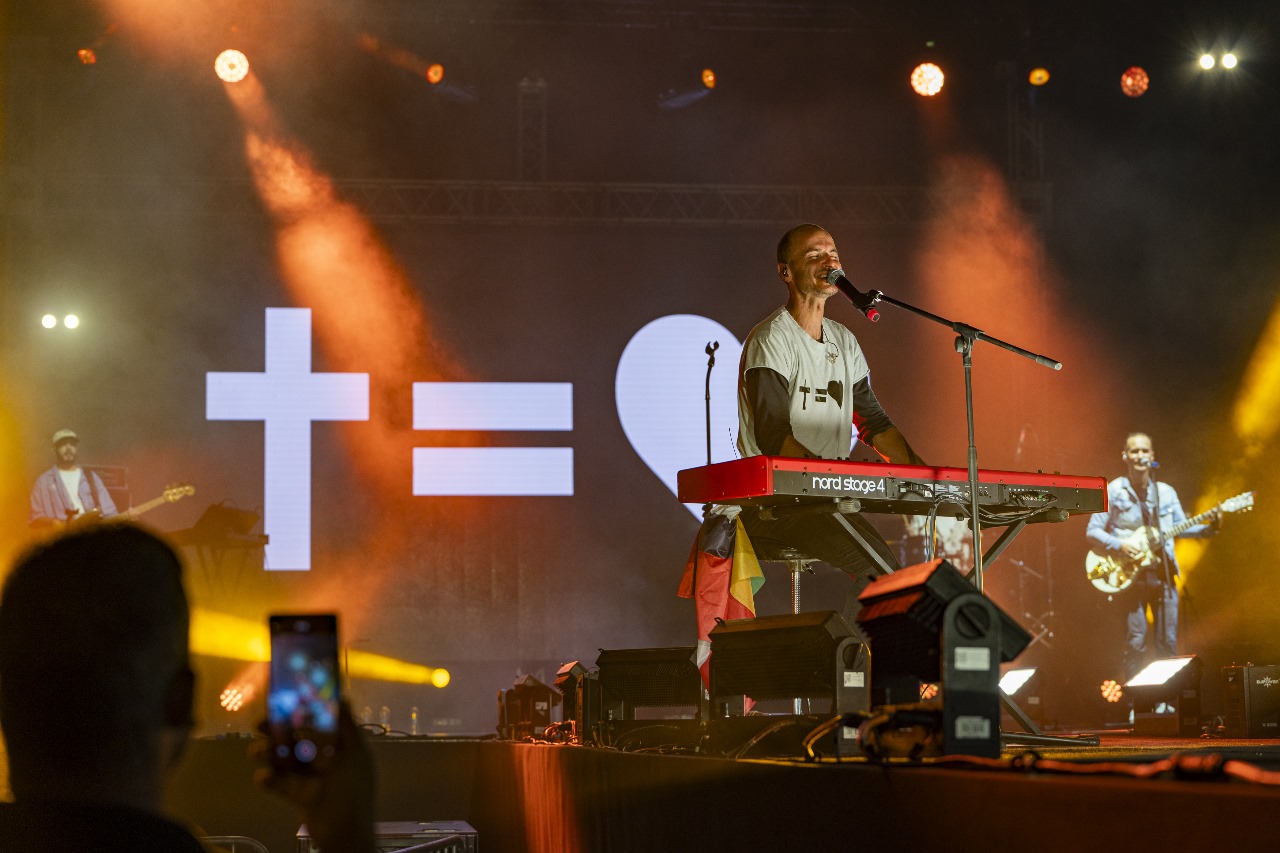 Musician playing keyboard and singing on stage with a band, backed by a large 'Cross = Heart' light display.