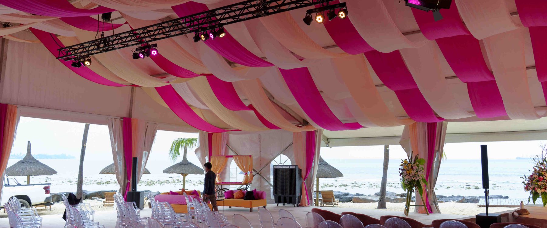 A large event tent by the beach is adorned with pink and white drapes and elegant lighting. Rows of transparent chairs face the ocean, creating a serene, festive atmosphere.