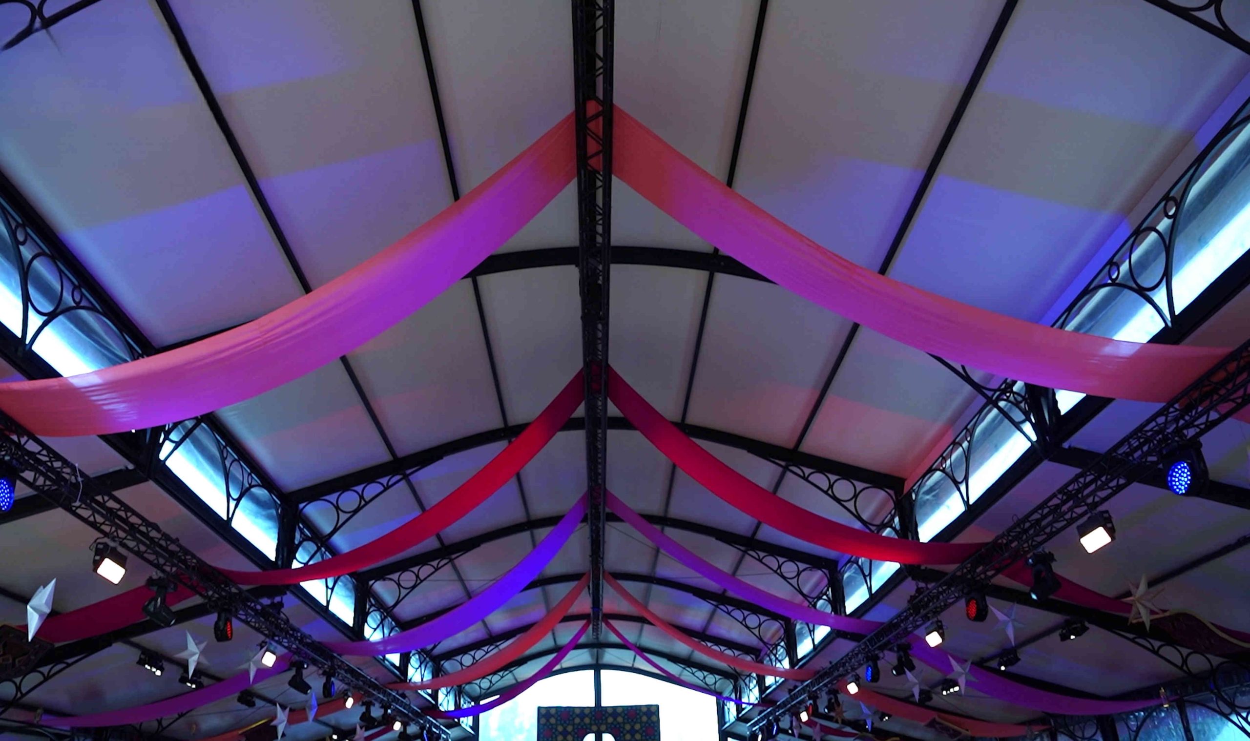 A ceiling view of a venue with elegant pink and purple draped fabric. Modern metal framework and soft lighting create a festive, lively atmosphere.