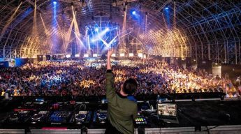 DJ performing in a vast, packed warehouse with arched ceiling. Vibrant lights and colorful beams illuminate the energetic crowd, creating a lively atmosphere.