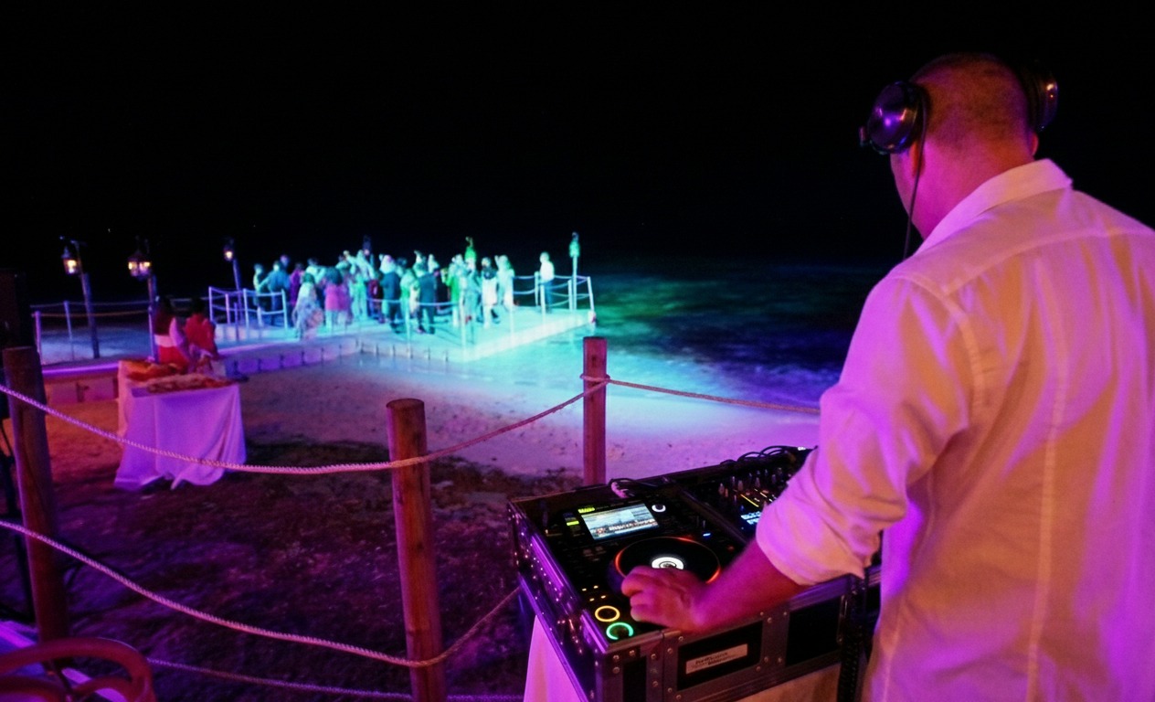 DJ in a white shirt mixes music under a starry night. Nearby, a lively group dances on a beach platform illuminated with colorful lights.