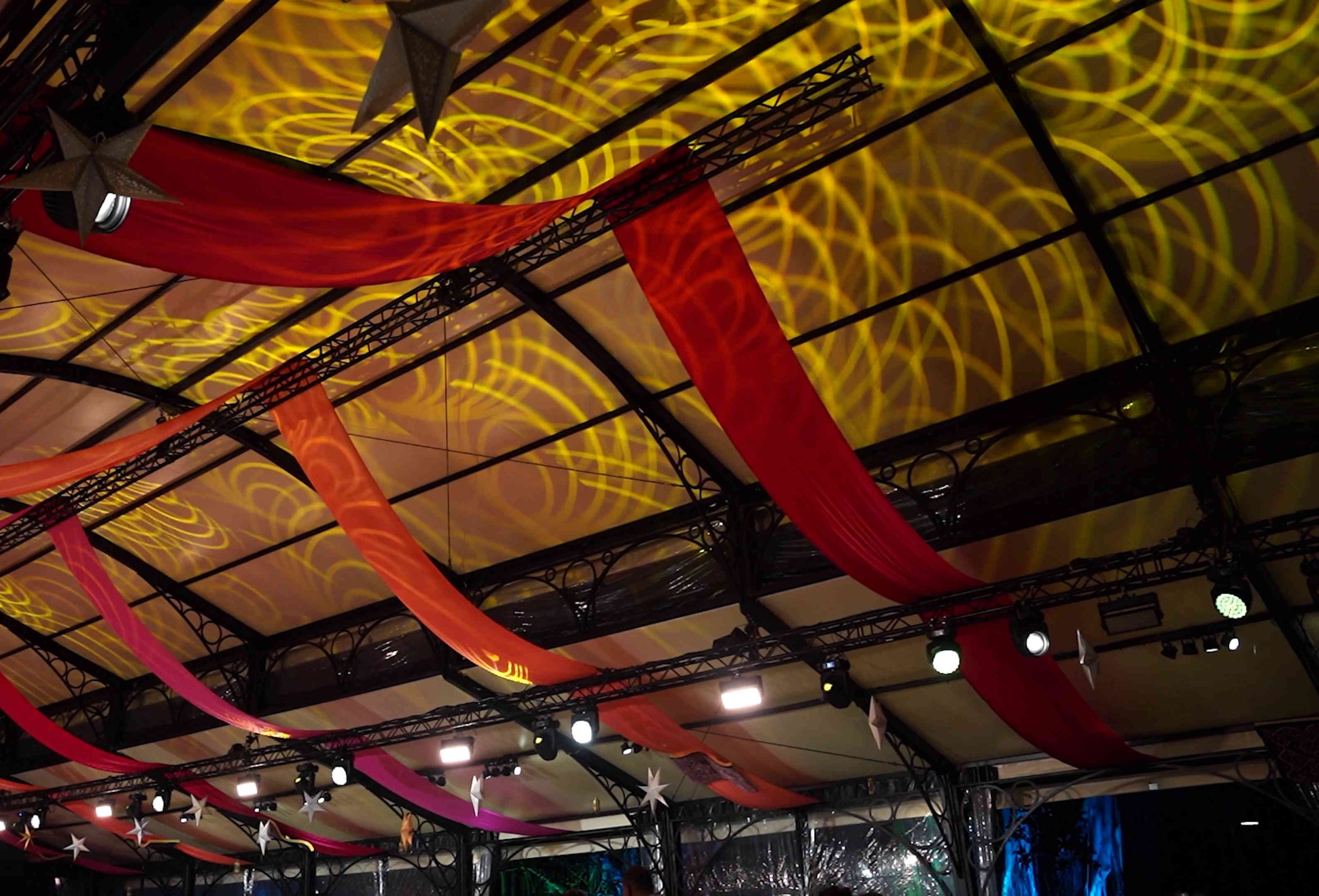 A colorful event ceiling with red and pink hanging ribbons and golden spiral light projections. The atmosphere is festive and vibrant, with spotlights below.