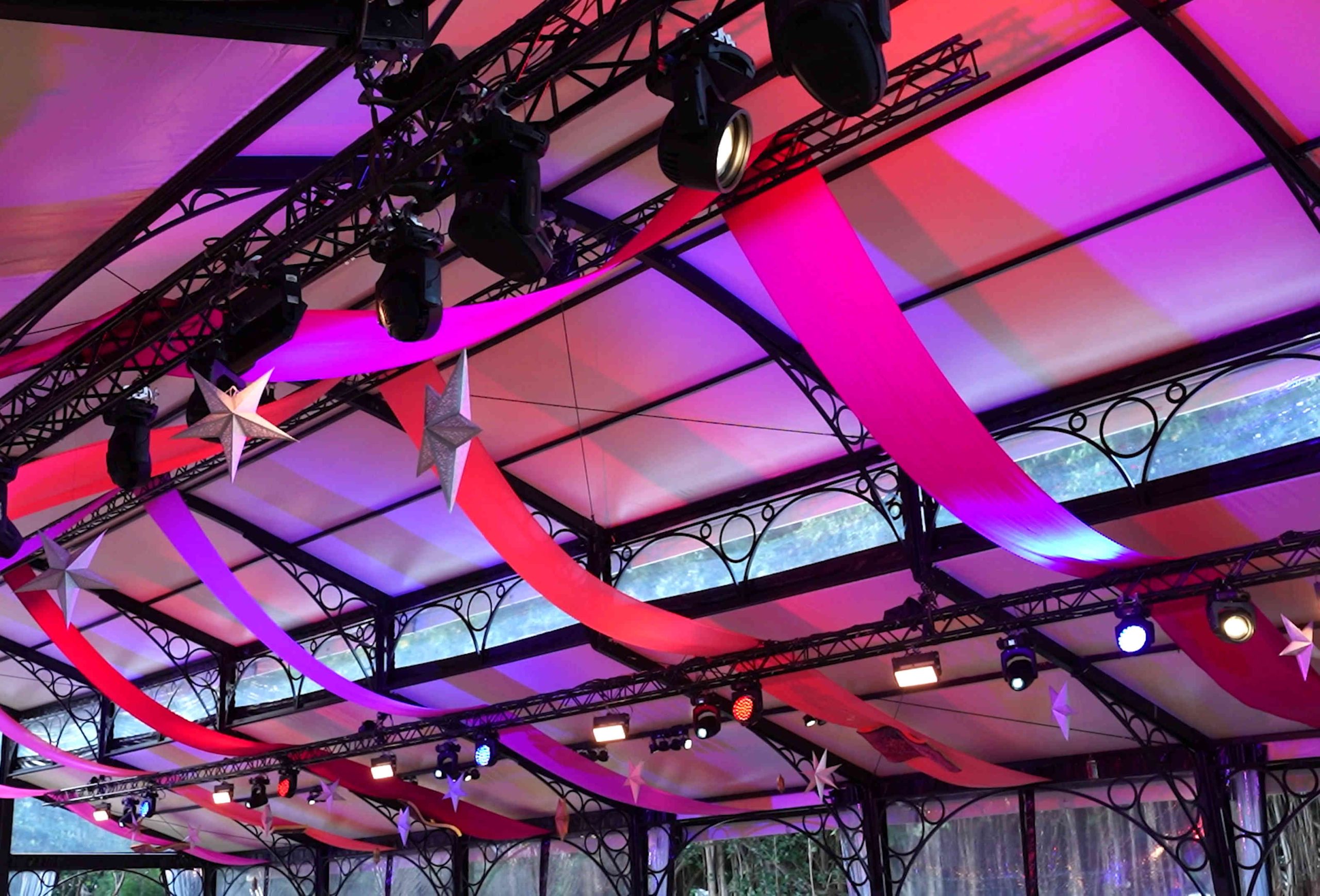 Event ceiling adorned with vibrant red and purple fabric drapes, illuminated by colorful lights, with hanging silver star decorations, creating a festive atmosphere.