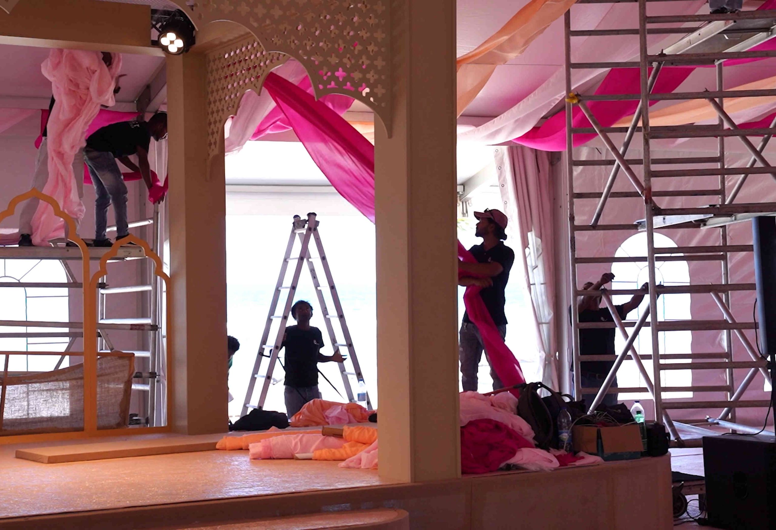 Event setup crew arranging vibrant pink and peach drapes on scaffolding inside a spacious venue with arched windows, creating an elegant atmosphere.