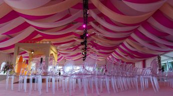 Opulent venue with pink and beige draped ceiling, clear chairs, and elegant floral arrangements, creating a festive and sophisticated atmosphere.