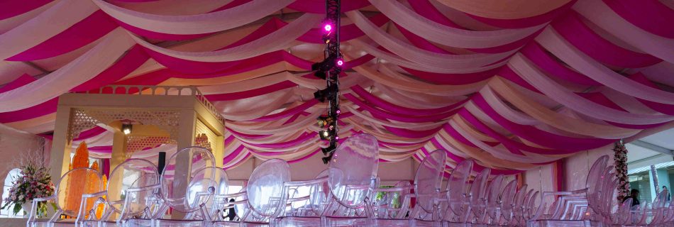Opulent venue with pink and beige draped ceiling, clear chairs, and elegant floral arrangements, creating a festive and sophisticated atmosphere.