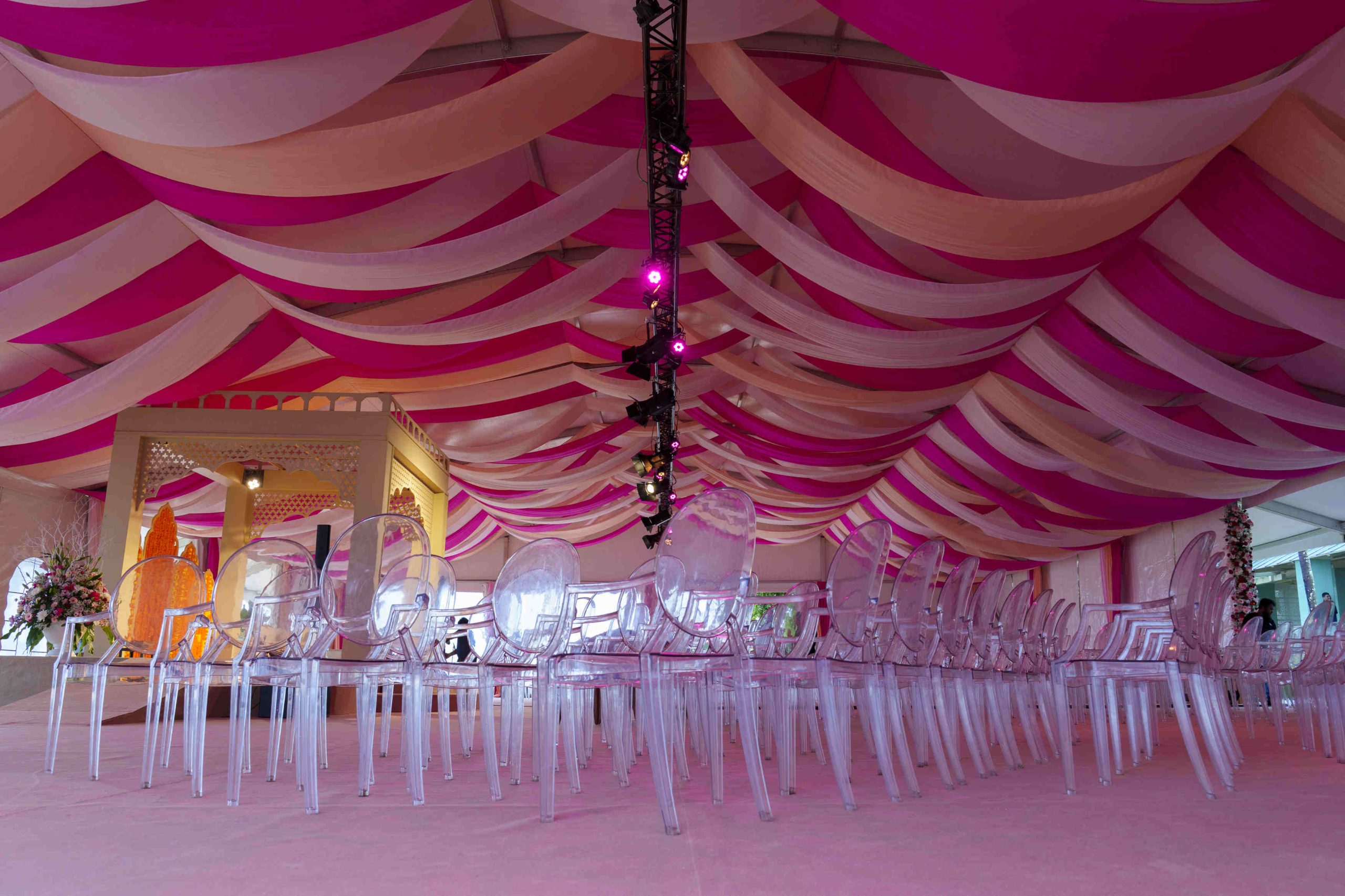 Opulent venue with pink and beige draped ceiling, clear chairs, and elegant floral arrangements, creating a festive and sophisticated atmosphere.