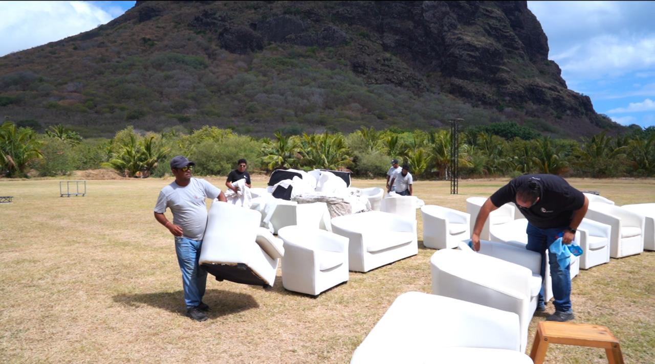 People arrange white lounge chairs on a grassy field under a clear blue sky. A mountain and palm trees form the scenic backdrop, conveying a relaxed, outdoor event setup.