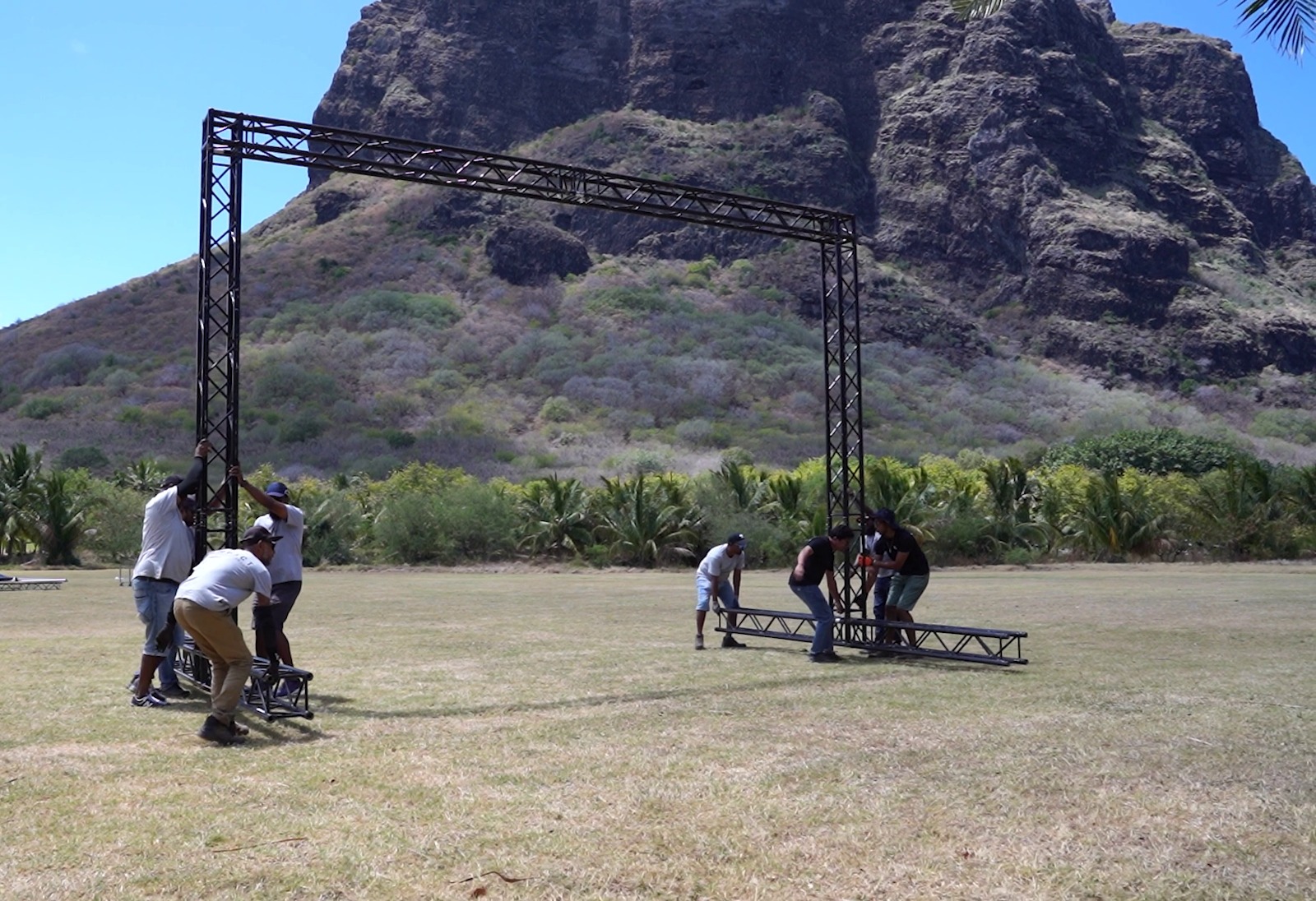 A group of people assemble a metal event structure in a grassy field under a large rocky mountain. Palm trees line the background, suggesting a tropical location.