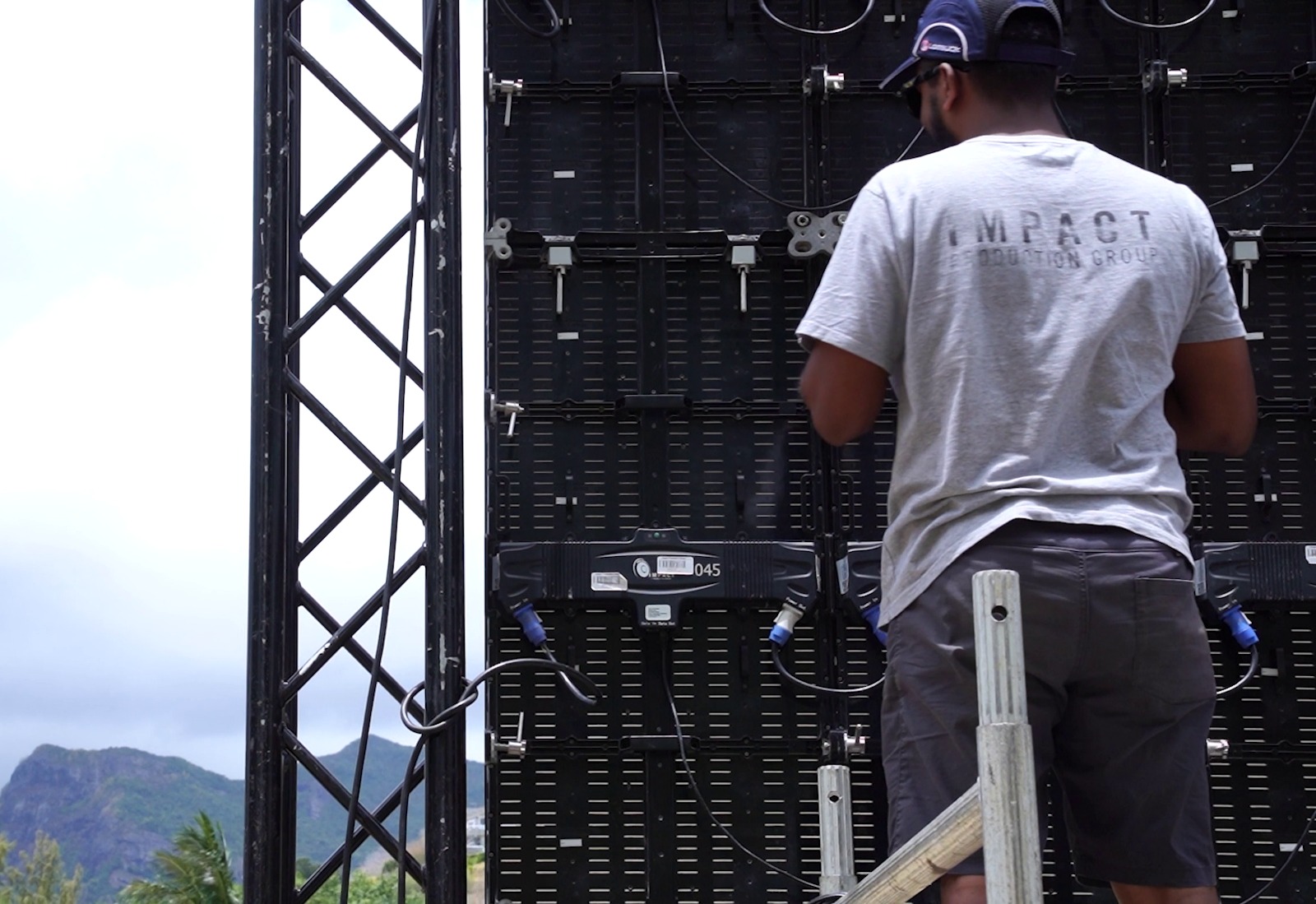 A person wearing a cap and a gray T-shirt stands on a ladder, adjusting a large back panel of electronic equipment outdoors. Mountains and trees are visible in the background.