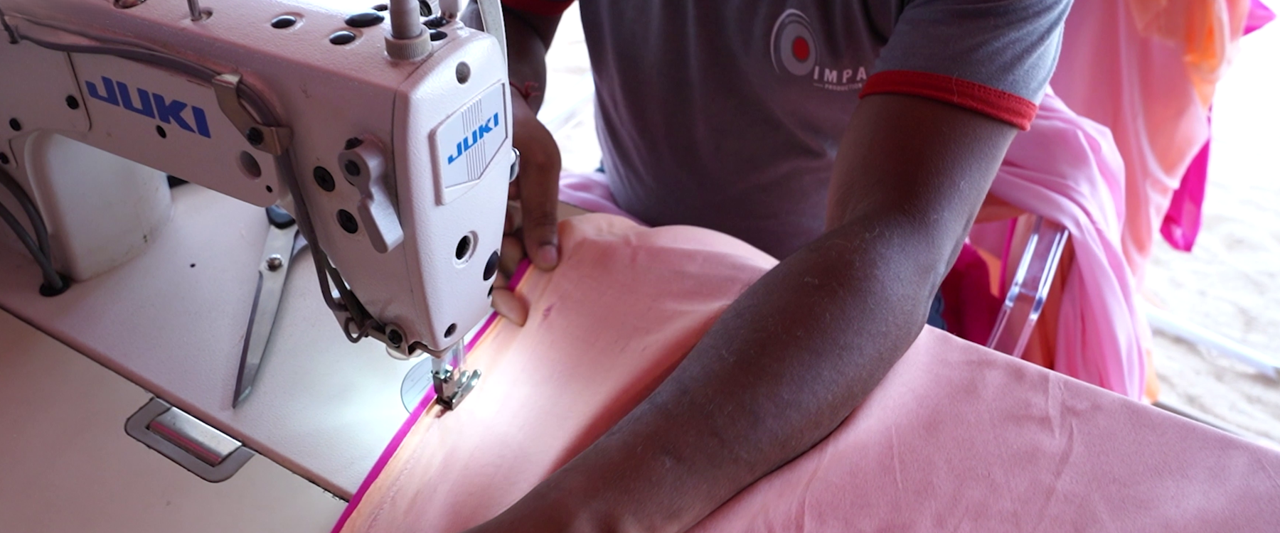 A person in a gray shirt operates a Juki sewing machine, stitching pink fabric. The scene conveys focus and skill in a textile work environment.