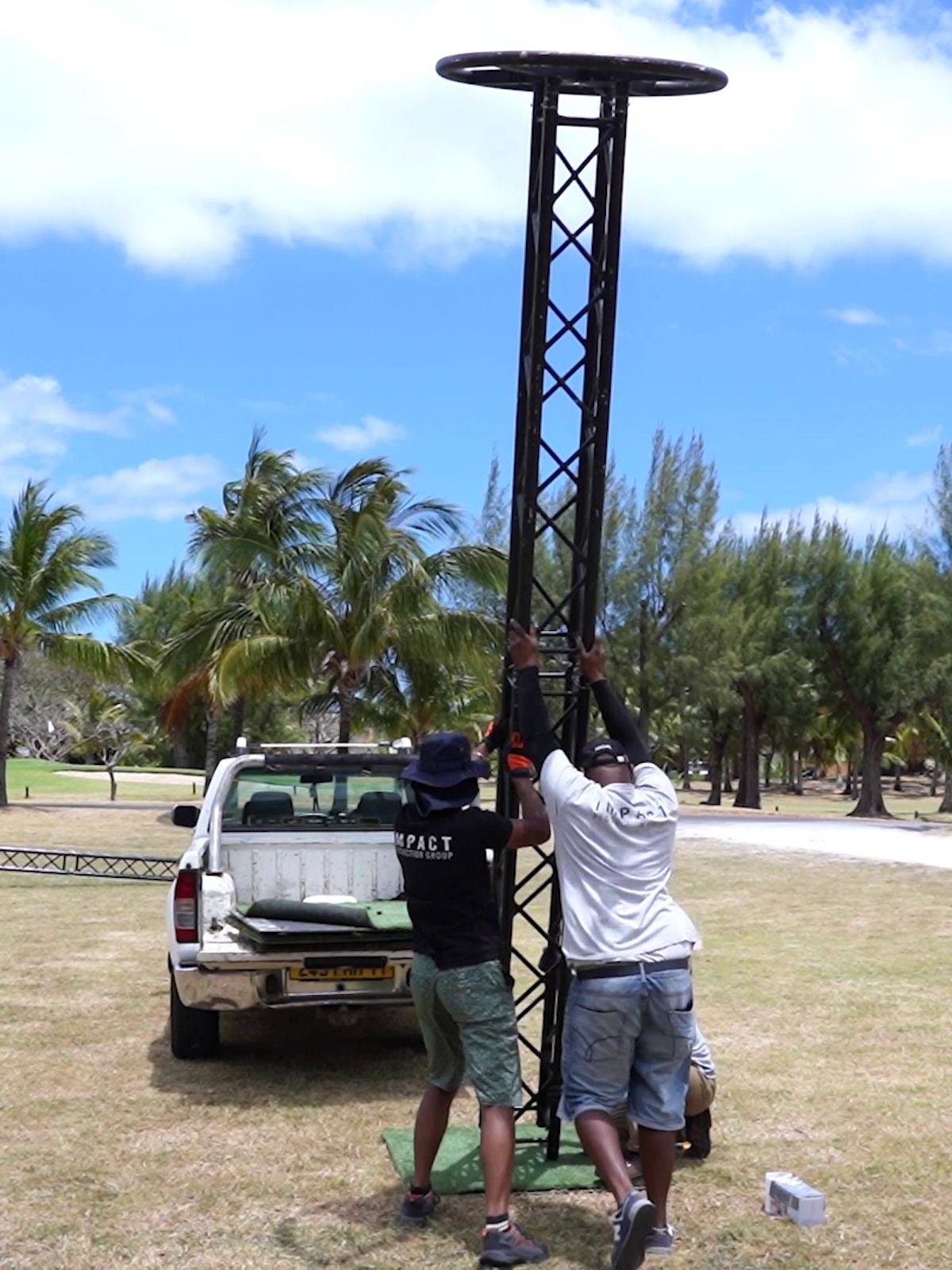 Two individuals are assembling a tall, metal tower on a grassy area near a white truck. Palm trees and a clear blue sky create a tropical setting.