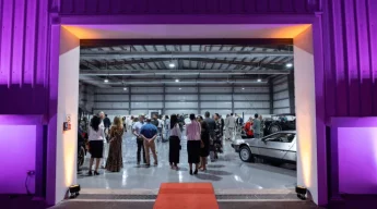 A large open space with a crowd of people mingling inside a hangar-like building. Classic cars are on display. The ambiance is elegant and upscale.