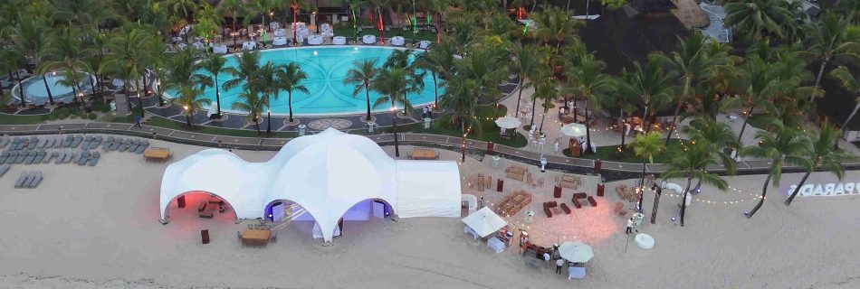 Aerial view of a beach resort with a large white tent glowing in red and blue on the sand. Nearby, a pool is surrounded by palm trees and festive lights.
