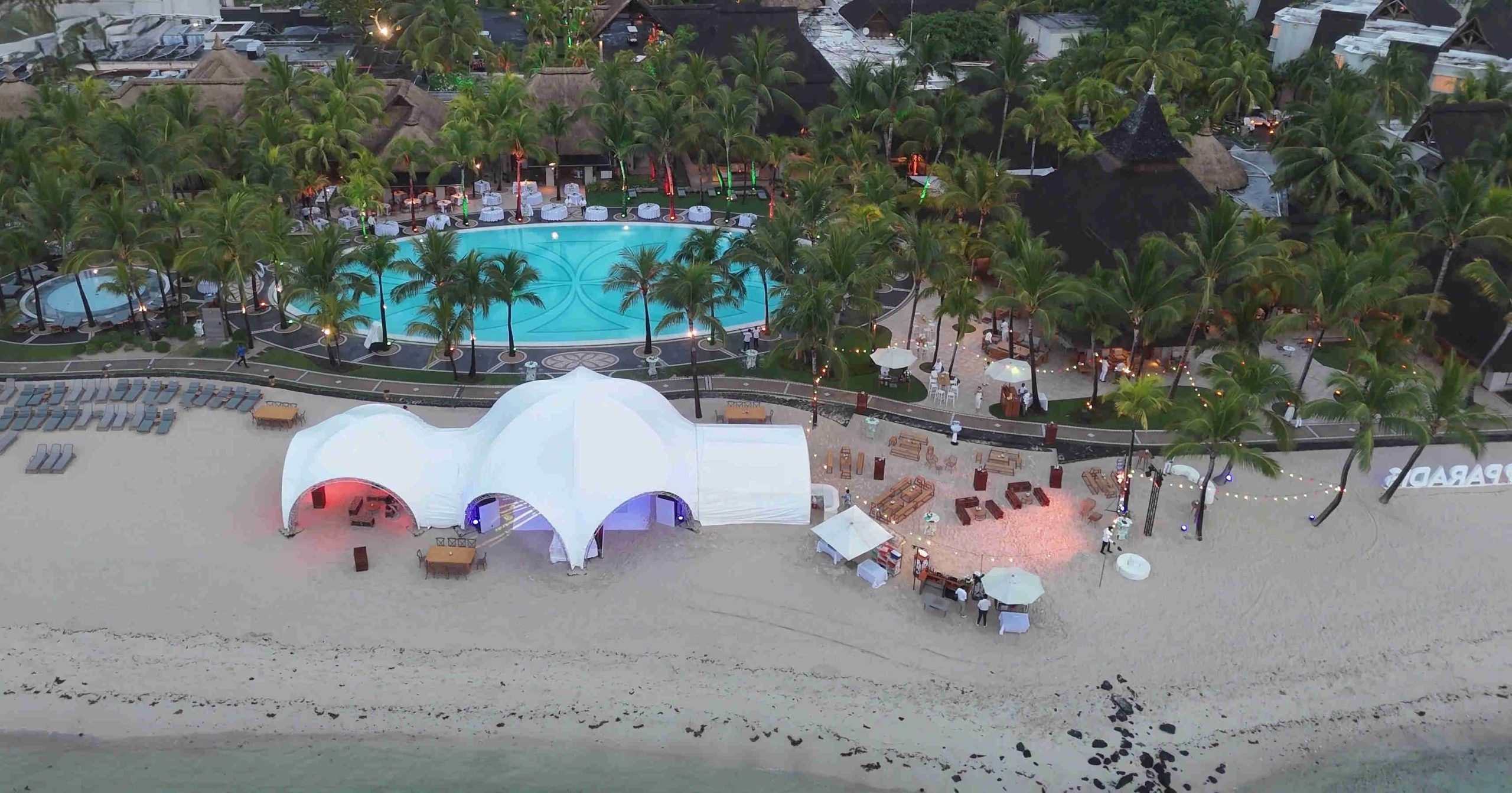 Aerial view of a beach resort with a large white tent glowing in red and blue on the sand. Nearby, a pool is surrounded by palm trees and festive lights.