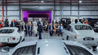 A man in a suit speaks to a seated audience at a car show, surrounded by classic cars in a spacious, well-lit showroom, creating an elegant atmosphere.