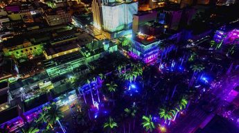 Aerial view of a vibrant cityscape at night, illuminated by colorful neon lights in blue, green, and purple tones, highlighting palm trees and buildings.